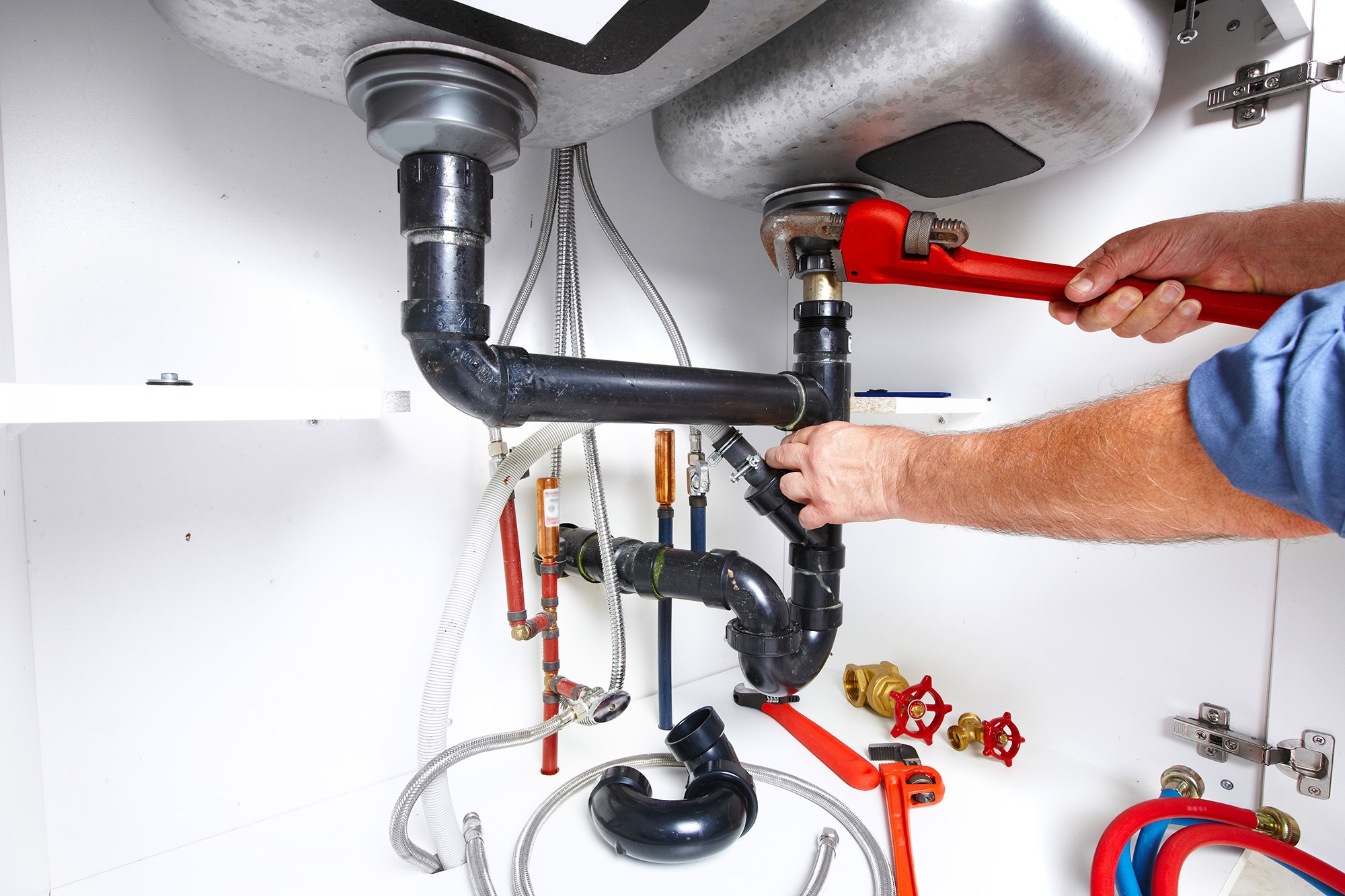 A person using a red pipe wrench to tighten plumbing pipes under a sink, illustrating DIY plumbing repairs and maintenance.