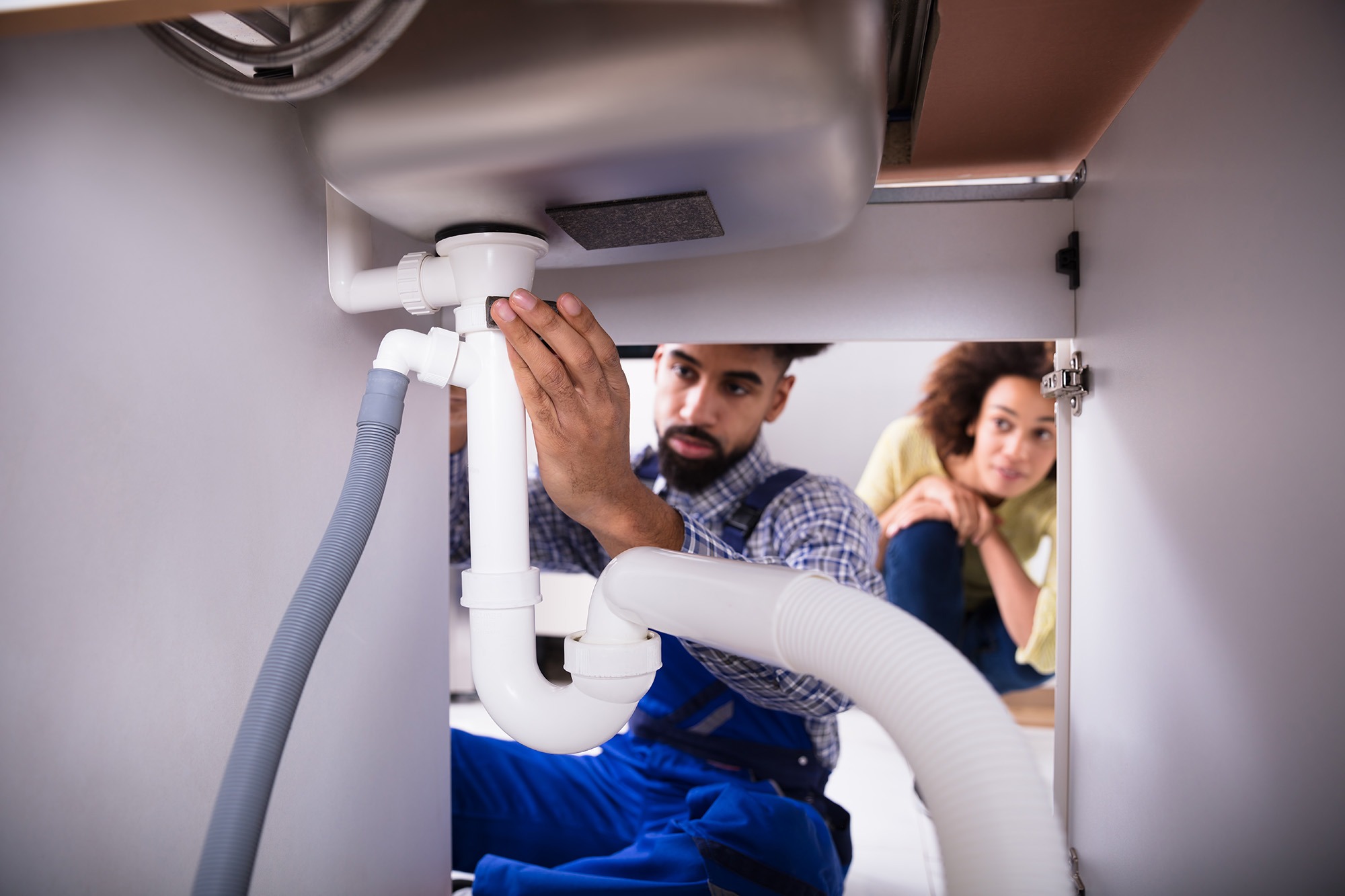 A plumber repairs a sink's plumbing under a countertop while an attentive woman observes, highlighting home maintenance and repair.