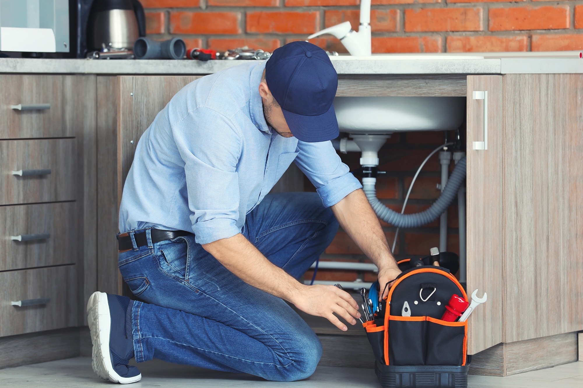 A plumber kneels by a kitchen sink, inspecting plumbing under a cabinet while accessing tools from a black and orange toolkit.