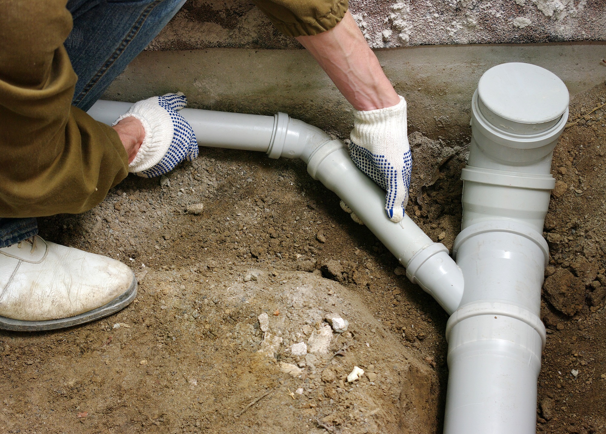 Person wearing gloves installs PVC pipes in a trench, demonstrating plumbing work related to drainage or sewage systems.