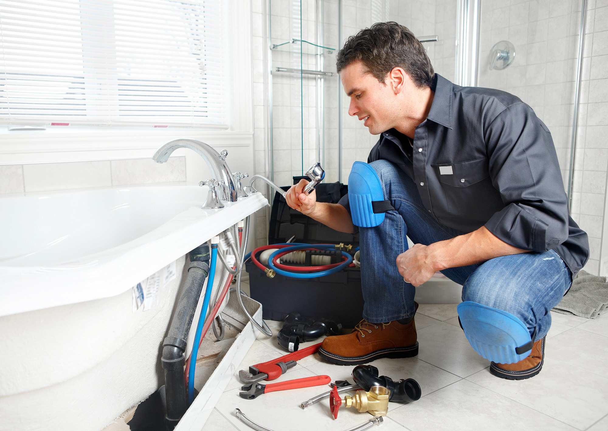 A plumber inspects a faucet installation under a bathtub, surrounded by tools and plumbing equipment, in a bright bathroom setting.