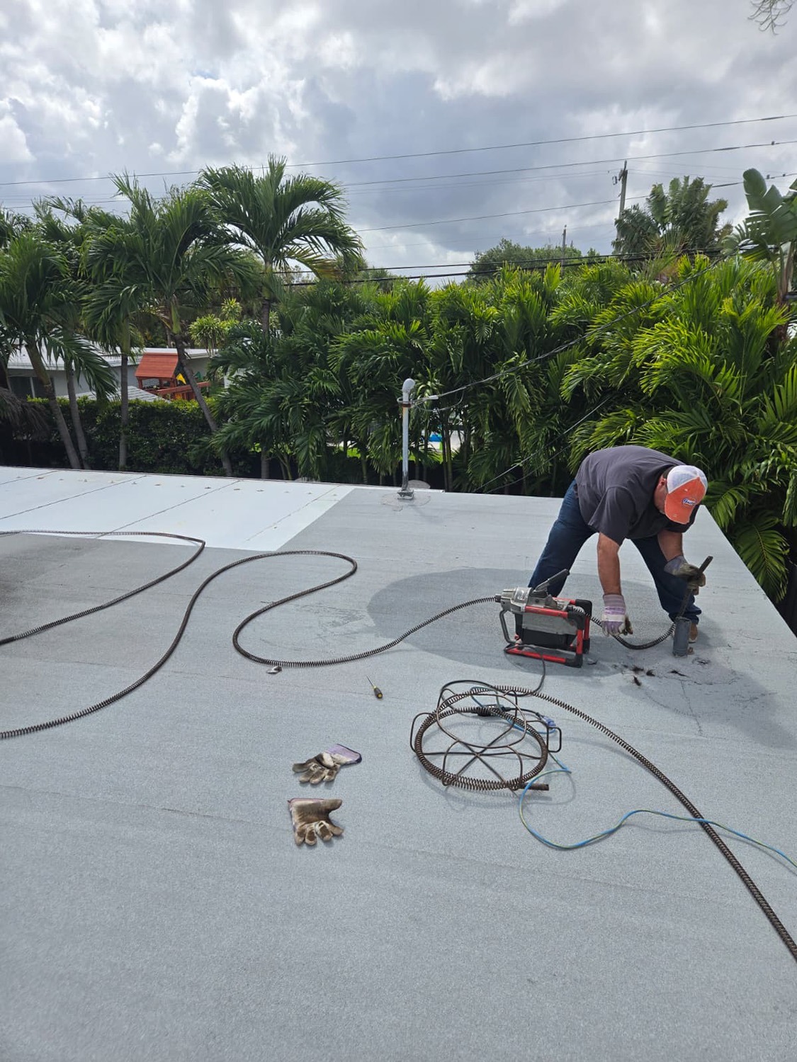 A worker repairs a flat roof, using tools and gloves lying nearby. Lush palm trees and a cloudy sky create a tropical backdrop.