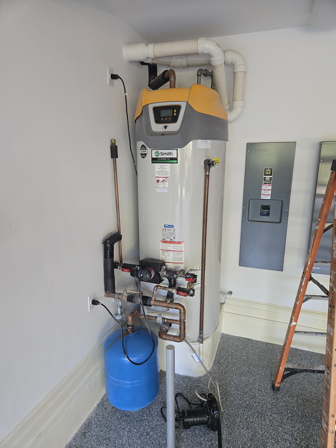 A modern water heater with a digital display is installed in a garage, connected to pipes and a blue pressure tank along with electrical and mechanical components.