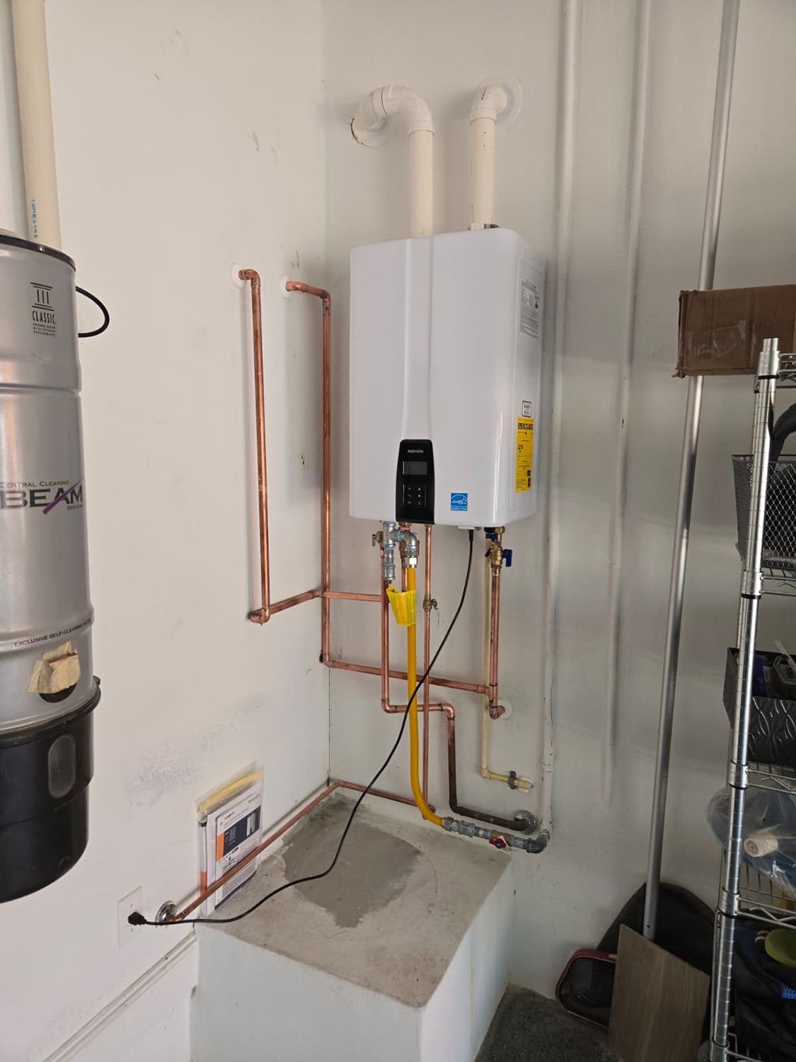 Tankless water heater mounted on the wall, connected to copper piping. A vacuum canister is nearby, with storage items to the right.