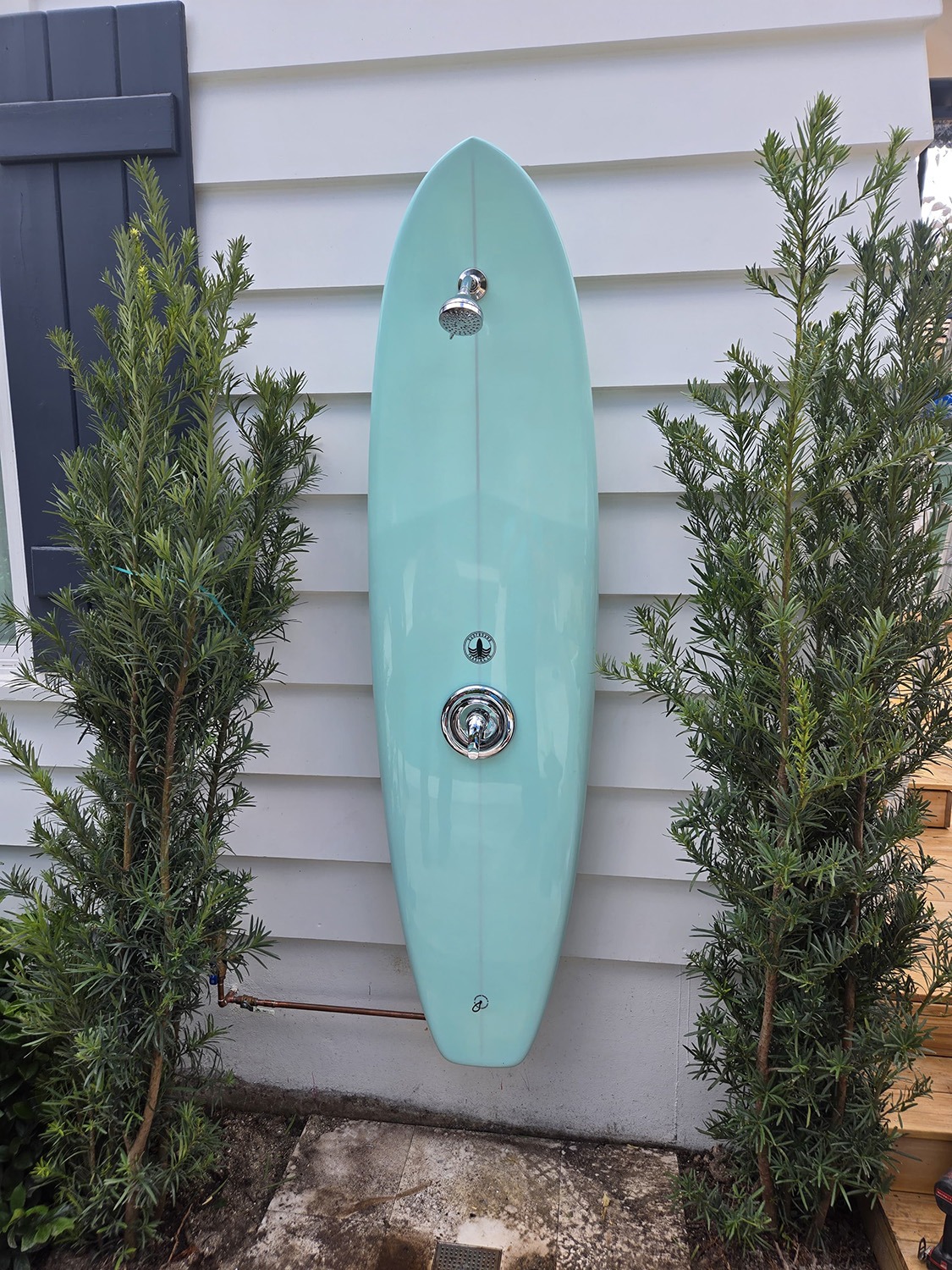 A teal surfboard-shaped outdoor shower mounted on a white wall, framed by green shrubs. Ideal for rinsing off after beach visits.