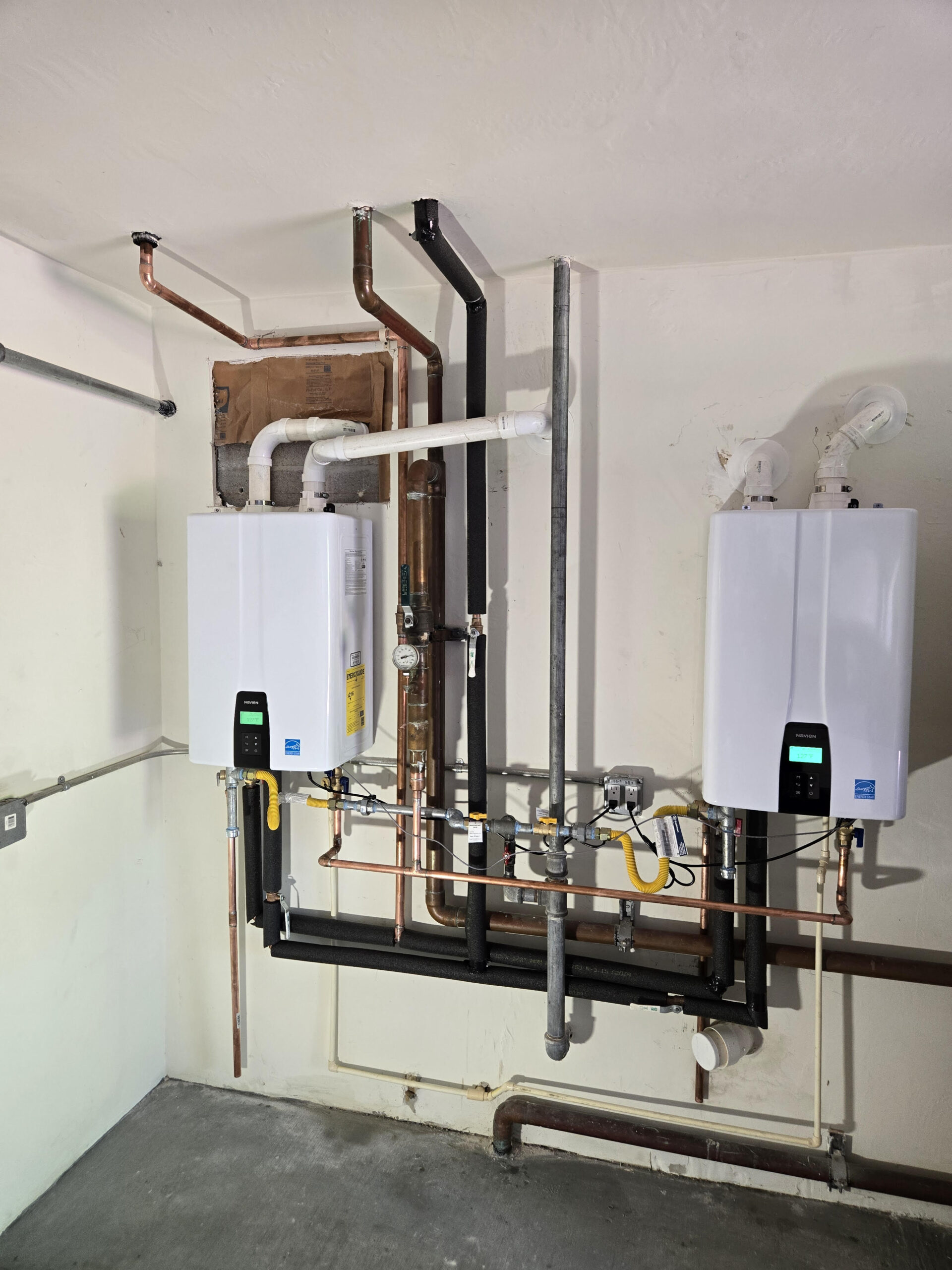 Two tankless water heaters mounted on a wall, with various copper and PVC pipes connecting them. This setup showcases an efficient heating system installation.