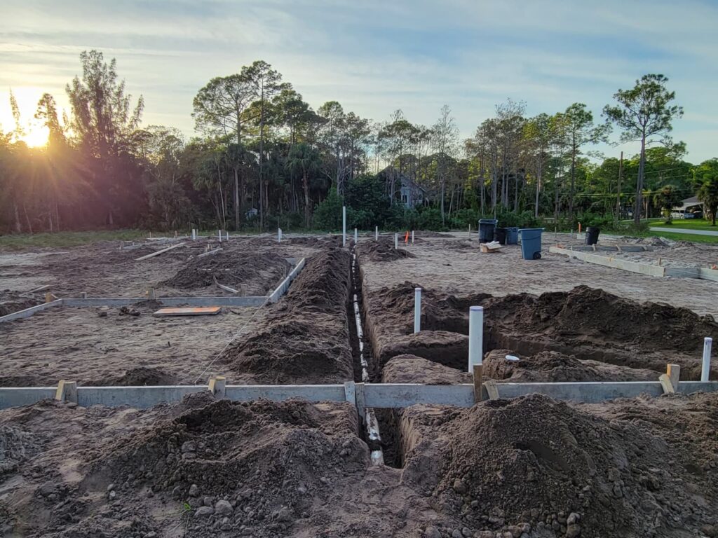Construction site with foundation trenches and PVC pipes. Framed in dirt, the area is surrounded by trees at sunrise, creating a calm, early morning scene.