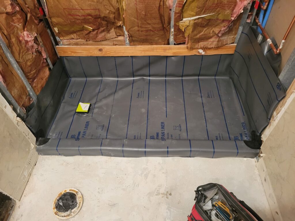 Shower construction in progress with exposed insulation and plumbing. A gray waterproof liner is laid on the floor, a toolbox and measuring tools nearby.
