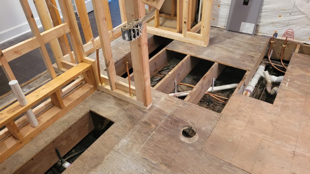 Exposed wooden framing and subflooring in a partially constructed room, revealing plumbing and wiring. The scene conveys construction progress.