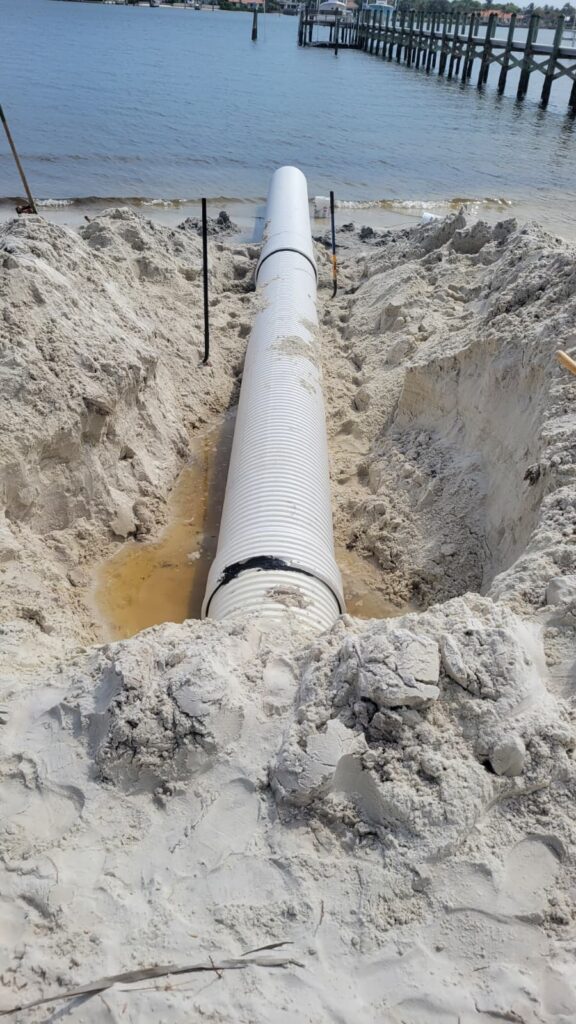 A large white drainage pipe partially buried in sand leads toward a serene body of water, with a pier extending out on the right under a clear sky.