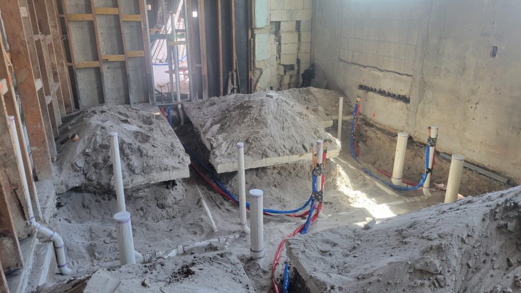 Interior construction site with exposed plumbing, showing dirt mounds and PVC pipes. The setting is industrial and in progress, with sunlight filtering in.