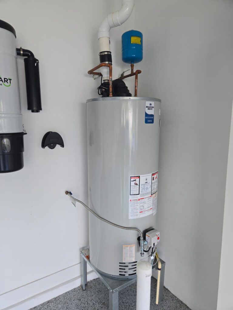 A vertical, silver water heater with labels stands in a white garage. It has copper pipes and a blue tank attached. The setting is clean and functional.